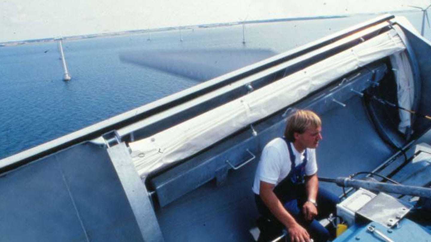 Worker in a white T-shirt and blue boiler suit sitting on top of a wind turbine.