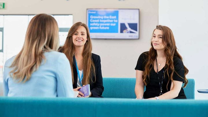 Three females in office attire sit on a blue sofa whilst talking.