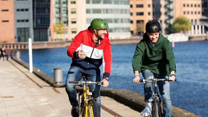 Two people wearing bike helmets cycling along the side of a river.