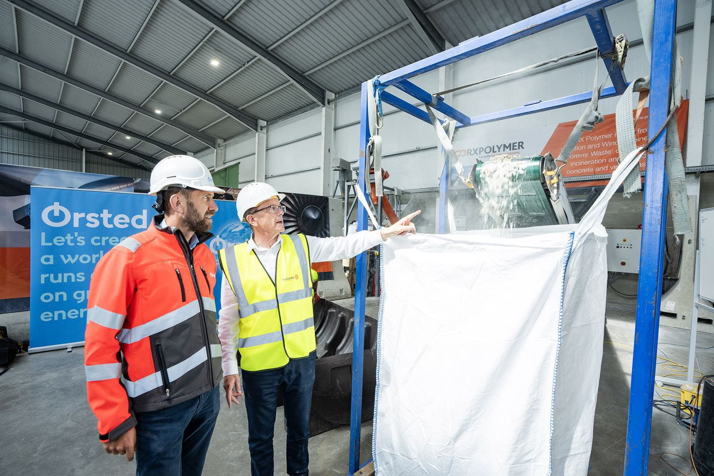 Two men watching a turbine blade being recycled in a factory setting.