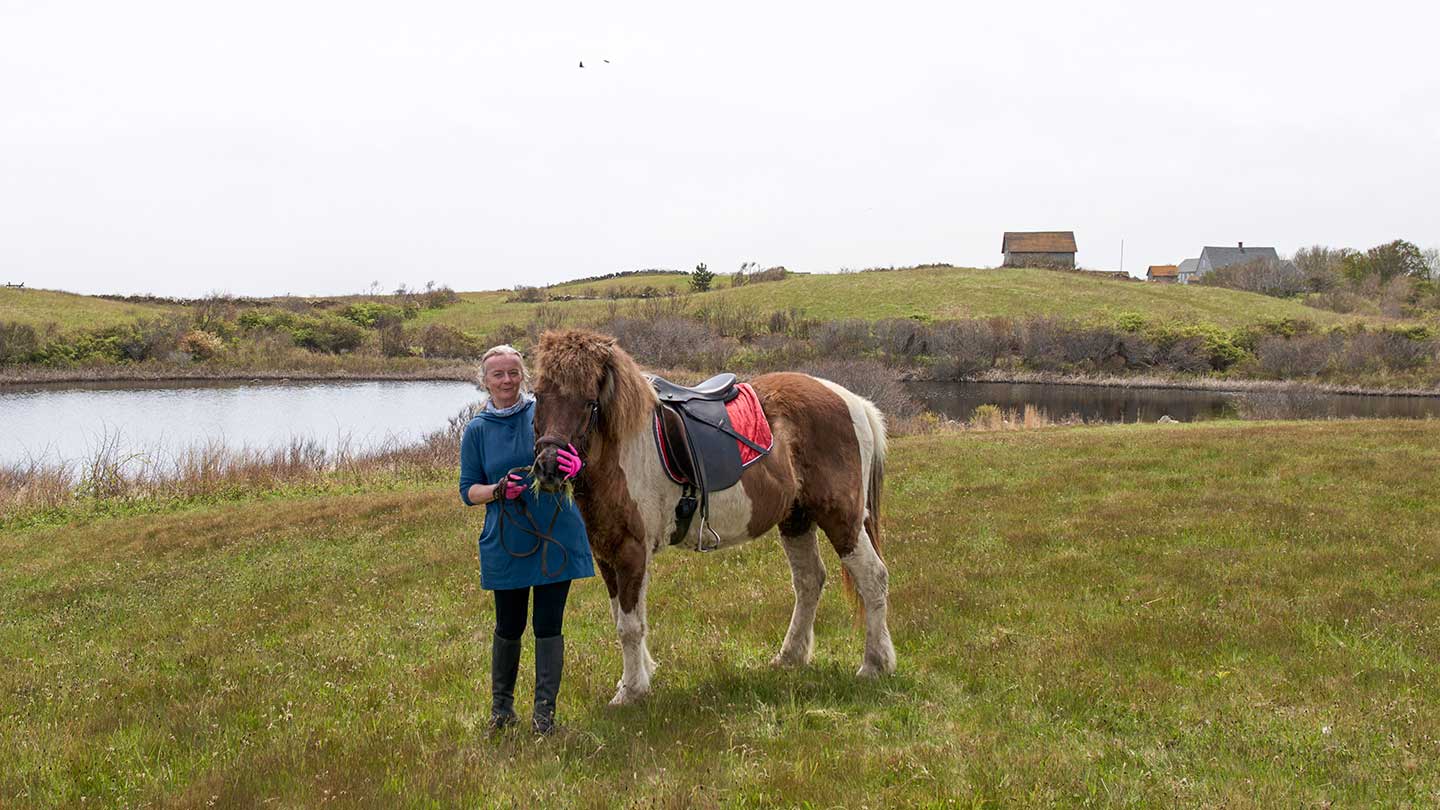 Dr. Susan Gibbons, Block Island local and science teacher, stands with her horse in a grassy field with a lake behind her.