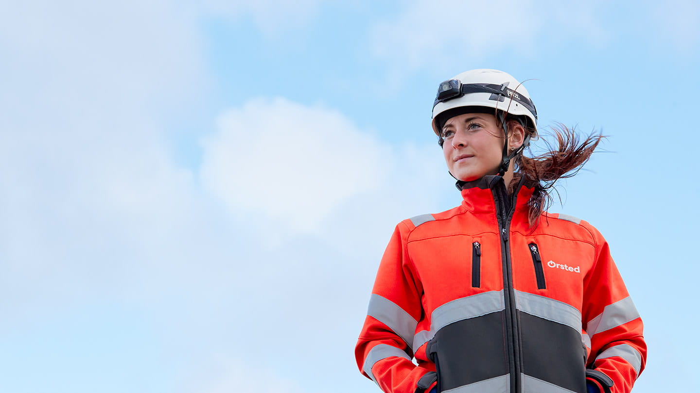 A female Ørsted employee wearing a hard hat and orange safety jacket, making a positive impact through clean energy.