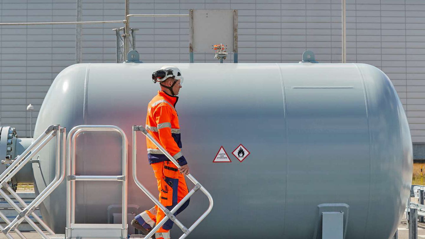 An Ørsted engineer in orange safety gear walking by a gray hydrogen tank, contributing the GreenFuels for Denmark project.