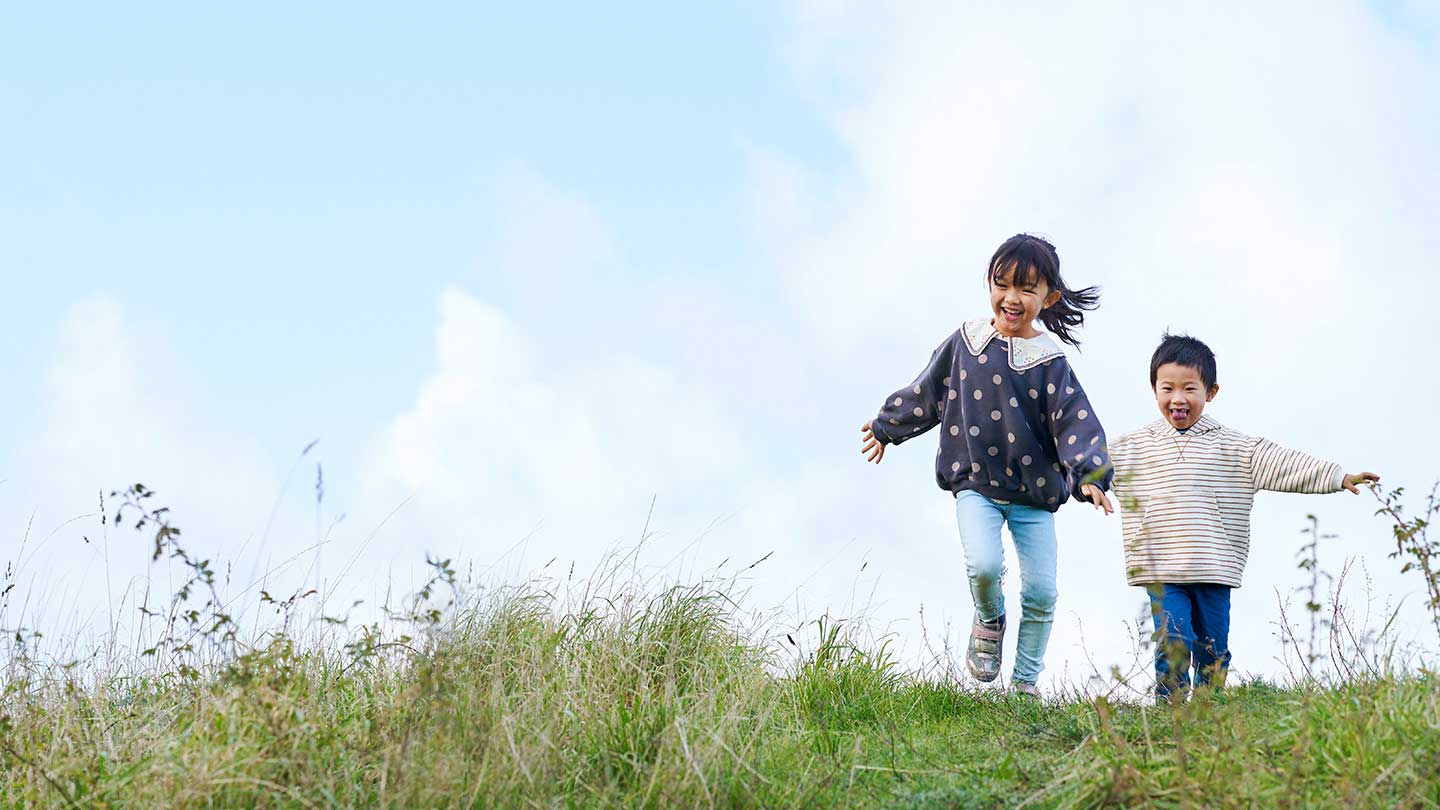 Two children, one wearing a dotted top and the other a striped hoodie, running in a green field.